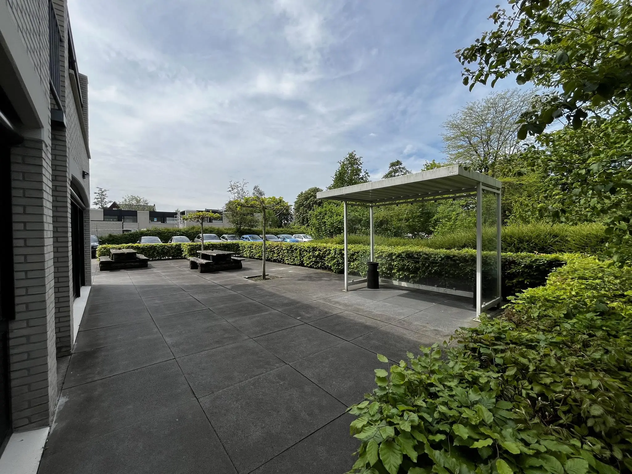 Moderne, betegelde buitenruimte aan de Burgemeester Verderlaan met zitbankjes, overkapping en groene beplanting.